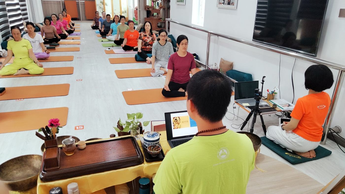 Một lớp yoga đang ngồi thiền trên thảm cam, người dạy quay lưng ở tiền cảnh